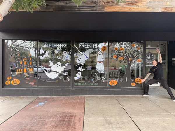 Halloween window mural by Andy Carlson at AE UNLTD Jiu Jitsu: a haunted house, ghosts wearing jiu jitsu belts, funny tombstones, and jack-o'-lanterns, with Andy posing beside it