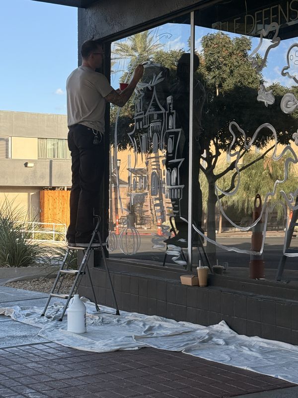 Andy Carlson on a step ladder painting the white outline of a haunted house on the AE UNLTD storefront window