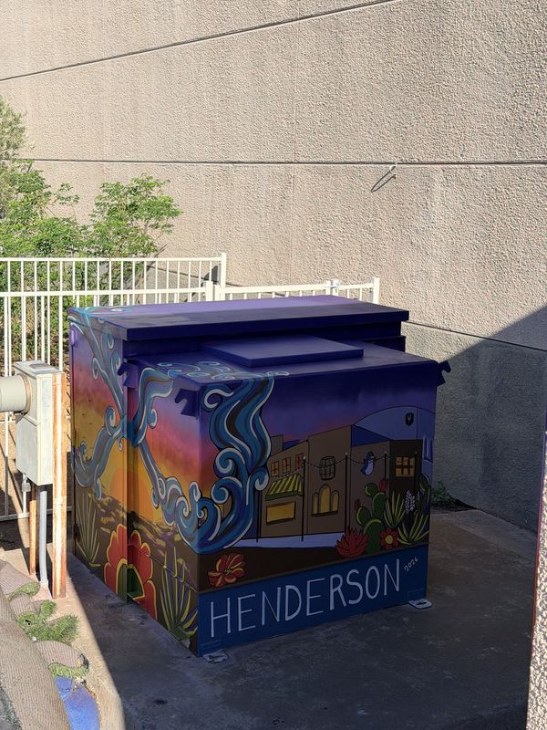 Utility box mural by Andy Carlson for the City of Henderson: a desert sunset, swirling blue water, downtown Henderson buildings with string lights, cactus and red flowers, and HENDERSON 2026 lettered along the base